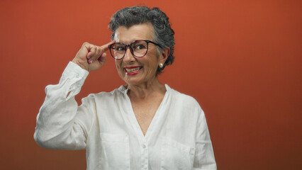 Obraz premium Senior woman with grey hair smiling and pointing to her head against an orange background, wearing glasses and a white shirt, conveying a thoughtful expression and confident attitude.