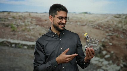 Young man in glasses holds a miniature shopping cart in a barren landscape implying consumerism...