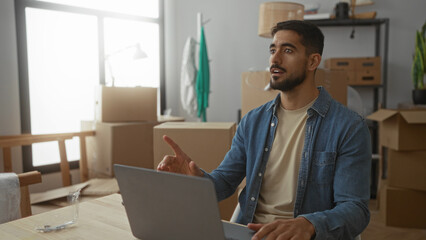 Young hispanic man in casual clothes works on laptop surrounded by moving boxes in new home living room setting.