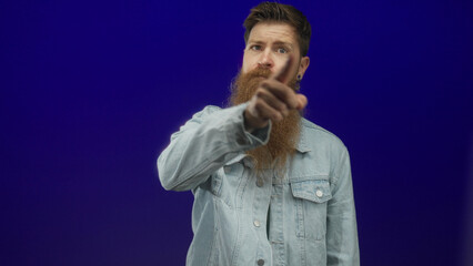 Young caucasian man with long red beard wearing denim jacket points finger toward corner in blue...