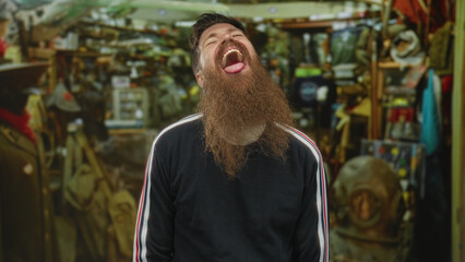 Fototapeta premium Young caucasian man with long beard sticks out tongue amid racks of tools in cluttered hardware shop building; amusement.