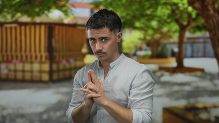 Young hispanic man gesturing with hands mimicking a gun in a sunny park setting surrounded by trees