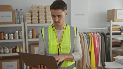 Young man in reflective vest working on a laptop in a charity donations center surrounded by...