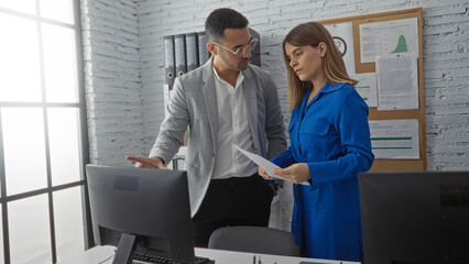 Man discussing work with woman in modern office setting with computers, documents on wall, and bright lighting.