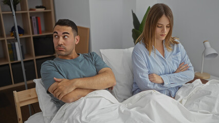 Man and woman sit in bed looking away from each other, expressing tension in a modern bedroom,...
