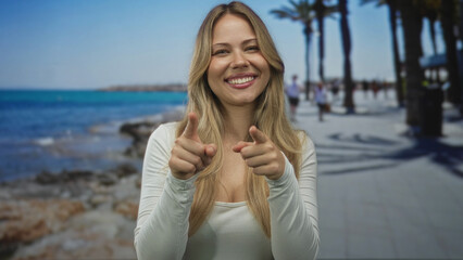 Blonde caucasian woman pointing finger guns at sunlit seaside promenade walkway; playfulness...