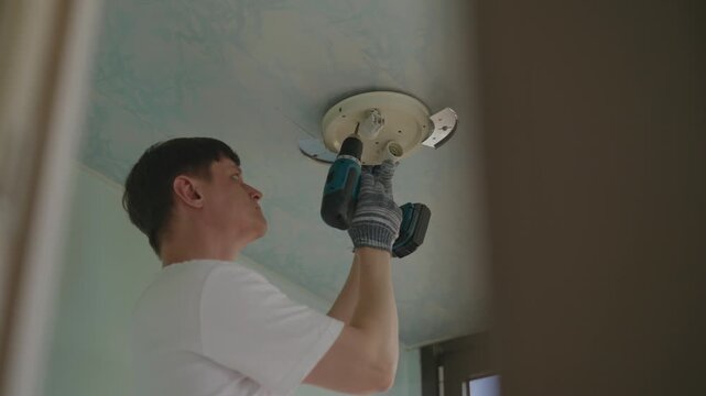 Shooting from below of electrician male in protective gloves installing ceiling light fixture using cordless screwdriver ensuring secure electrical connections in living room. Shooting in slow motion.
