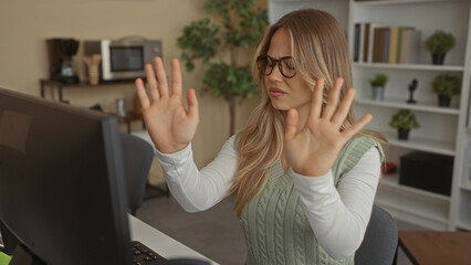 Woman with blonde hair wearing glasses hands raised with palms out at computer desk in office;...