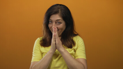 Woman praying hands gesture in studio with an orange wall, wearing a yellow shirt with a subtle...