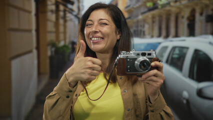 Middle aged hispanic woman smiling and holding vintage camera shows thumbs up on city street near...