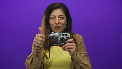 Middle aged hispanic woman holds vintage film camera and gives thumbs up in studio; confidence...