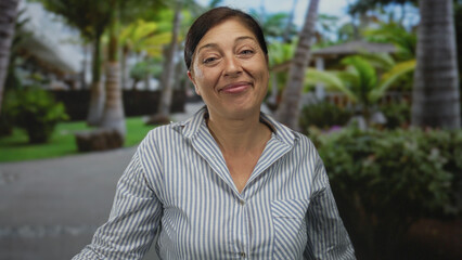 Fototapeta premium Middle aged hispanic woman smiling in lush green park under palm trees on sunny day wearing striped shirt; serenity.