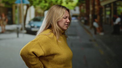 Woman grimacing with back pain on urban street dressed in yellow sweater, highlighting discomfort,...