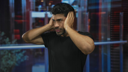 Man pressing hands over ears in hotel building with a tense expression and visible muscle...