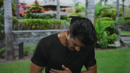 Young man holding stomach with hand while bending forward in green park outdoors; digestive...