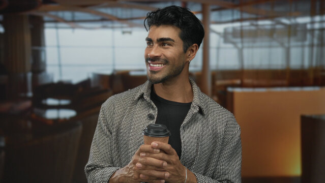 Young hispanic man smiling while holding paper coffee cup and enjoying morning beverage in modern hotel lobby; relaxation.