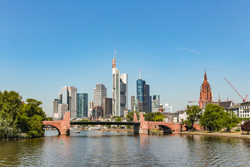 Obraz premium A Frankfurt city skyline in Germany with a bridge over a Main river. The bridge is red and the water is calm. Against the background of the blue sky.