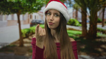 Woman in santa hat smiles and beckons with hand in street setting under green trees; friendly...