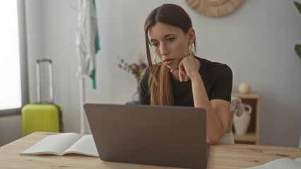 Woman holds pen to forehead with hand on chin while working on laptop at desk in building; focused...
