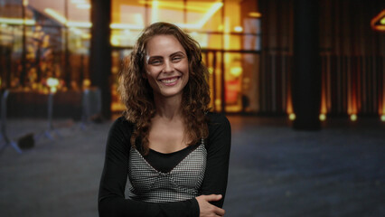 Woman smiling confidently on city street with warm lighting in the background, exuding candid energy and happiness amid urban evening ambiance outdoors.