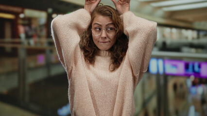 Woman expressing surprise indoors at a mall with her hands on head wearing glasses and a sweater...