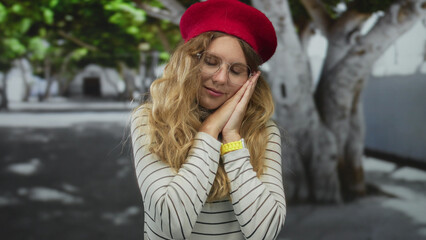Woman with hands resting in a courtyard features blonde young hair under a red beret and glasses in...