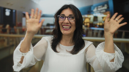 Smiling woman waves hand in bustling mall center with blurred shoppers and railings behind her;...