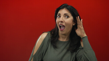 Woman cupping her right ear and leaning forward in studio with bright red background to hear faint...
