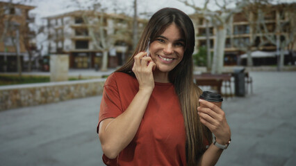Young hispanic woman talking on smartphone while holding coffee cup outdoors on city street with...