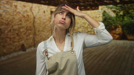 Young blonde woman in apron flicks bare hand with sideways glance on wooden patio; indifference...