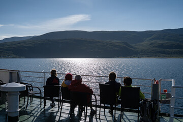 Group of people sitting on the ferry deck and enjoying the view on the fjord and green norwegian coast during summer season.