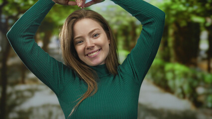 Woman in green sweater smiling and raising hands above head to form heart shape in forest pathway...