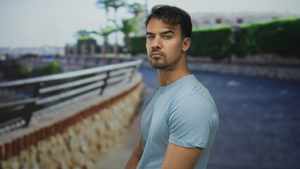 Young hispanic man standing outdoors by a fence in casual attire on a sunny day.