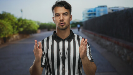Young hispanic man in referee uniform stands on a city street, outdoor setting highlighted by greenery, conveying urban sports vibe and professional determination.