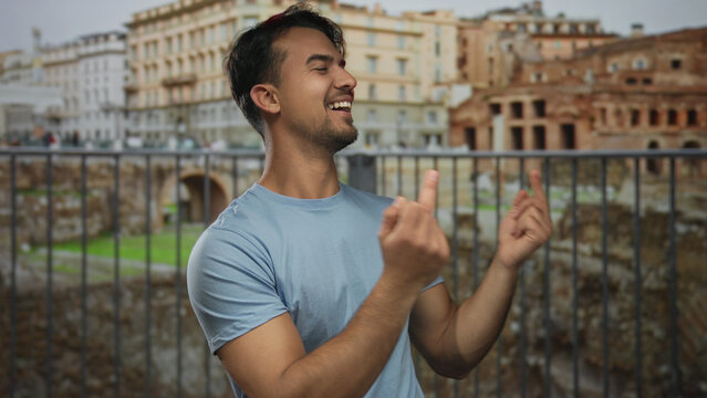 Hispanic man making gestures with middle fingers while laughing in front of roman ruins in an outdoor city setting conveys humor and irreverence.