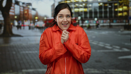 Fototapeta premium Woman in bright orange prison jumpsuit stands on busy urban street, expressing emotion outdoors with blurred city background in early evening light.