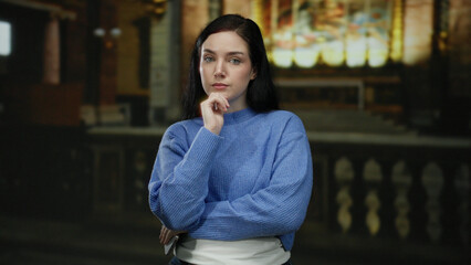 Woman in blue sweater standing thoughtfully in serene church setting, with soft light illuminating...