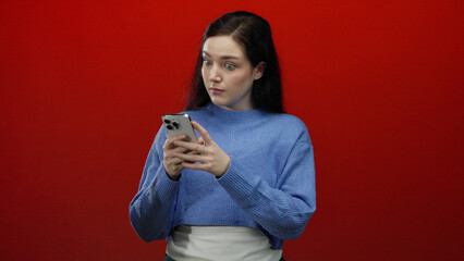 Woman excitedly reacts to smartphone in front of vibrant red background displaying joyful surprise.