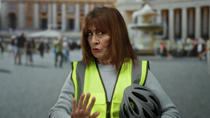 Woman in reflective vest holding helmet extends hand towards camera in busy square in vatican city,...