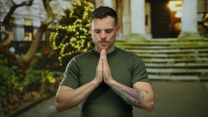 Young man in green shirt stands on urban street with hands in prayer gesture, lights glowing in background, serene expression, tattoo on arm, evening time, peaceful pose outdoors.