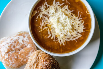 A Bowl Of Traditional French Onion Soup Topped With Grated Cheese And White Bread Rolls Topped With Flour And Seeds