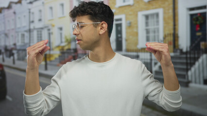 Young hispanic man wearing glasses gestures with both hands while speaking on a busy city street;...