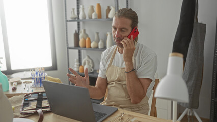 Man holds smartphone to ear while sitting at pottery studio desk with laptop and brushes;...