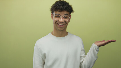 Young hispanic man wearing glasses and white sweater smiling holds open palm in green studio;...