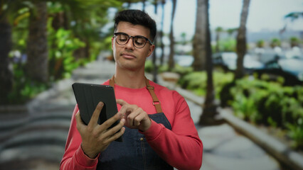Obraz premium Young man in glasses using tablet outdoors in a lush park setting, wearing casual red attire and apron, looking focused and engaging with the touchscreen device.