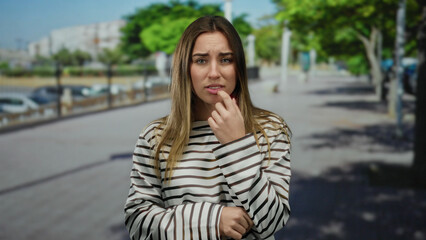 Woman wearing striped shirt looking concerned standing on street outdoors in sunny urban area with...