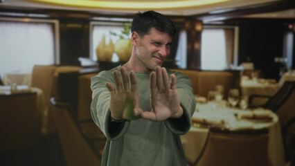 Hispanic young man holds both hands forward for stop gesture while standing by restaurant table;...