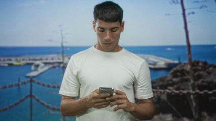 Man taps smartphone while standing on a weathered dock at a bustling port by the seaside in bright...