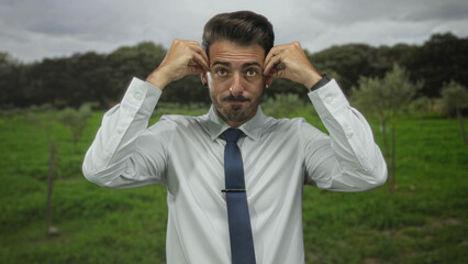 Man in crisp white shirt and navy tie raises arms toward sky and furrows brows in lush forest...