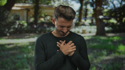 Young hispanic man clutching chest in lush sunlit park clearing during daytime, wearing green...
